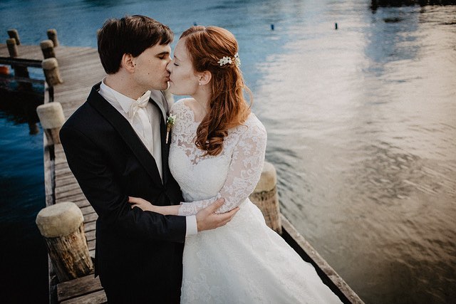 A bride and groom share a loving kiss on a wooden pier, creating a romantic wedding moment by the water.