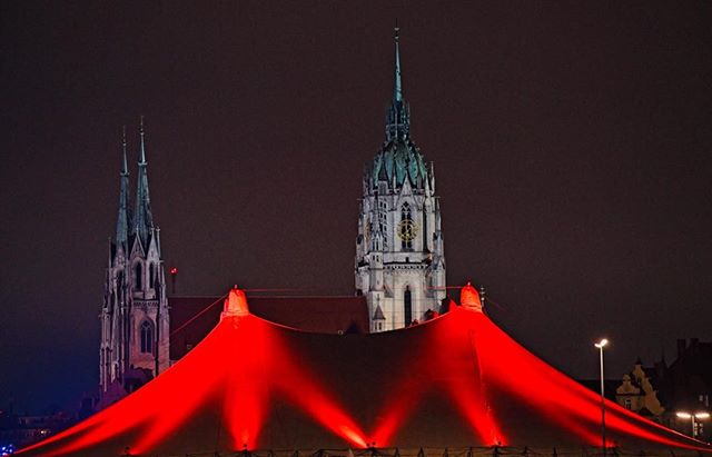 Illuminated church towers rise above a red-lit tent in a city at night. 