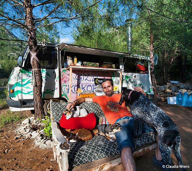 A man relaxes on a sofa with his pets in front of a camper van in a forest setting.