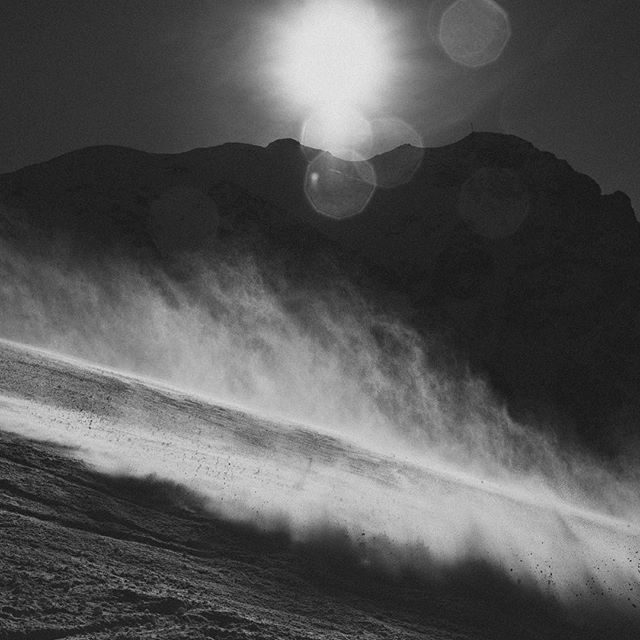 A monochrome landscape depicting a snowy mountain with strong winds blowing snow across the surface.
