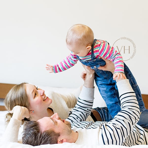 A happy family plays together, with the father lifting the baby up as the mother smiles on the bed.