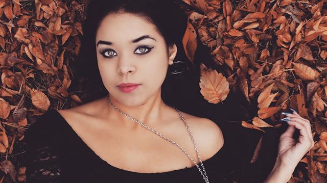 A woman lies amongst autumn leaves wearing a black top and a silver necklace. 