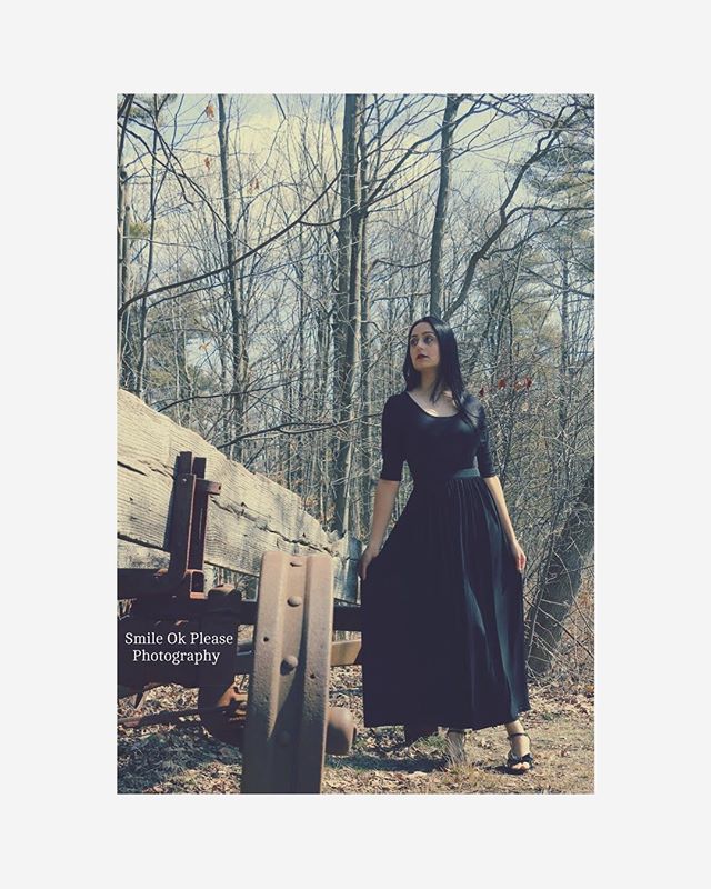 A woman in a black dress poses in a wooded area by an old rusty wheel for a fashion shoot.