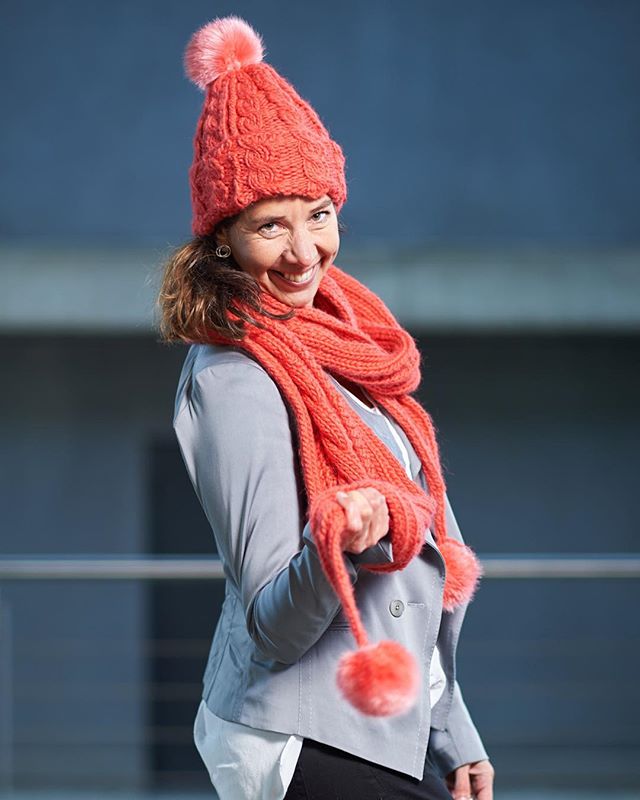 A cheerful woman smiles wearing a coral hat and scarf with a gray jacket in an outdoor setting.