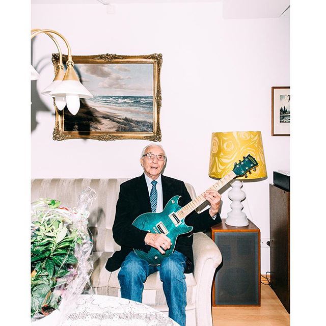 An elderly man sits on a couch playing a teal electric guitar in a well-lit living room.