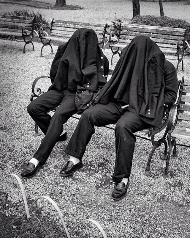 Two figures sit on a park bench, draped in coats concealing their identities in a black and white image.