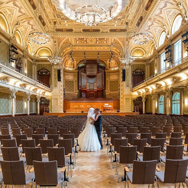 A bride and groom embrace in a grand, ornate concert hall before their wedding ceremony.
