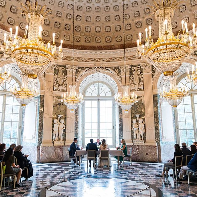 A wedding ceremony takes place in a grand ballroom, with the bride and groom at a table surrounded by guests and opulent chandeliers.