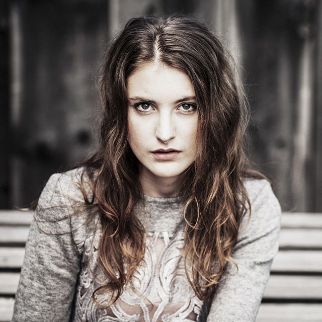 A young woman with long brown hair and a septum piercing poses for a portrait outdoors.