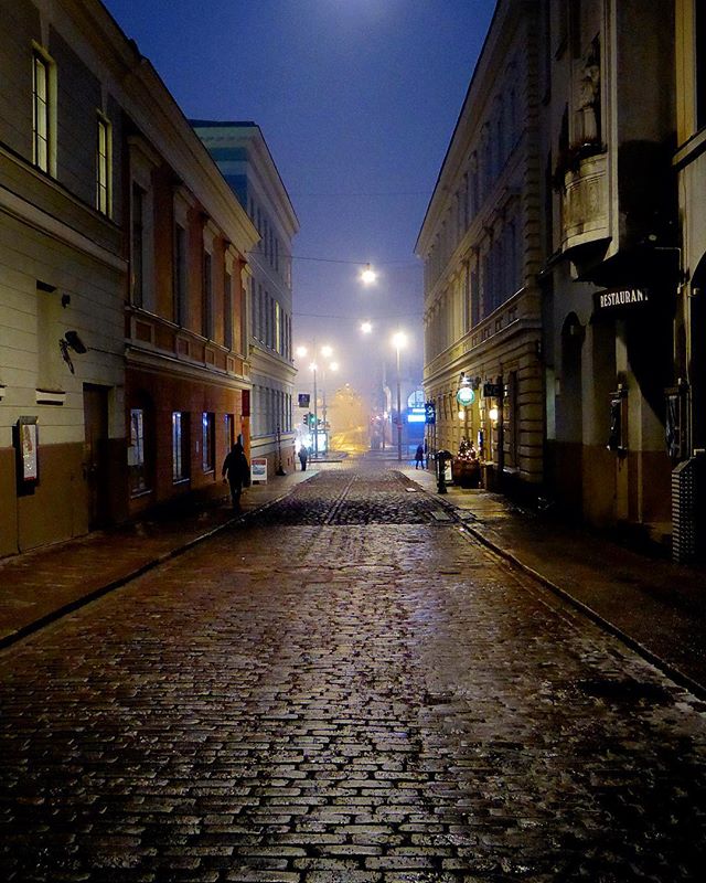 A foggy city street at night lined with buildings and people walking. The scene is atmospheric and moody.