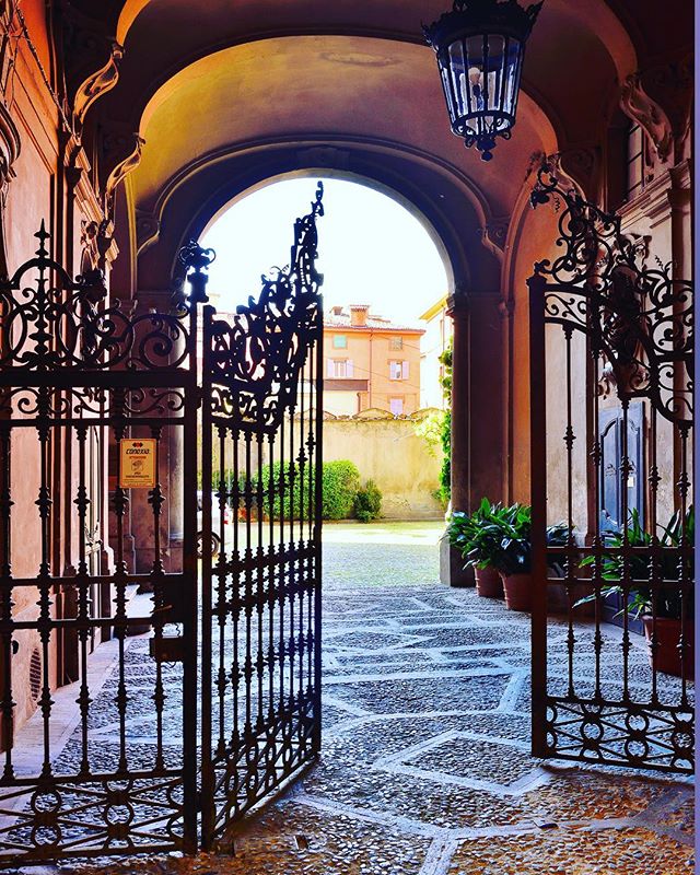 An ornate gate opens to a charming courtyard with stone paving under an arched passage in Europe.