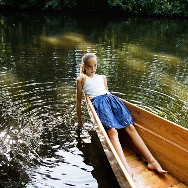 A young girl enjoys a calm day on the water, sitting in a wooden boat surrounded by a lush green landscape.