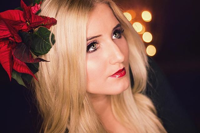 Elegant woman with blonde hair and a poinsettia flower in her hair, wearing red lipstick in a glamorous portrait.