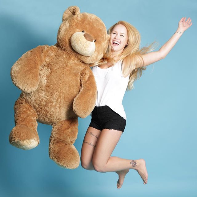 A cheerful woman jumps with joy beside a large teddy bear against a bright blue backdrop.