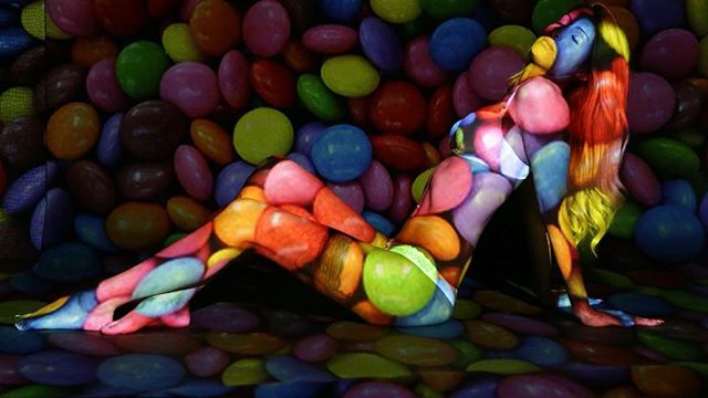 A model showcases vibrant candy-themed body painting in a creative, surreal studio art shot.