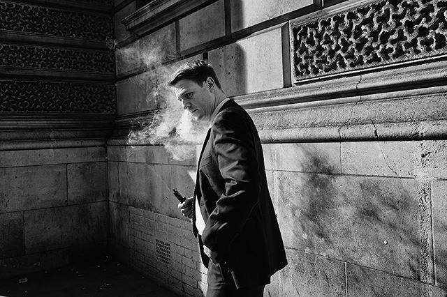 A man in a suit stands on a city street, exhaling vapor from an e-cigarette in a black and white image.