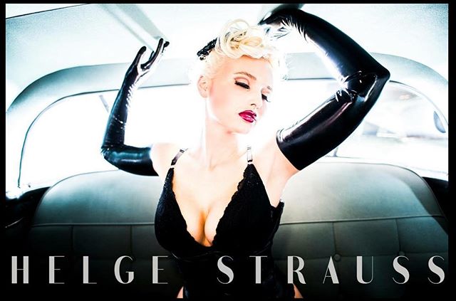 A vintage-style portrait of a blonde woman in a car, wearing latex gloves and an elegant black dress.