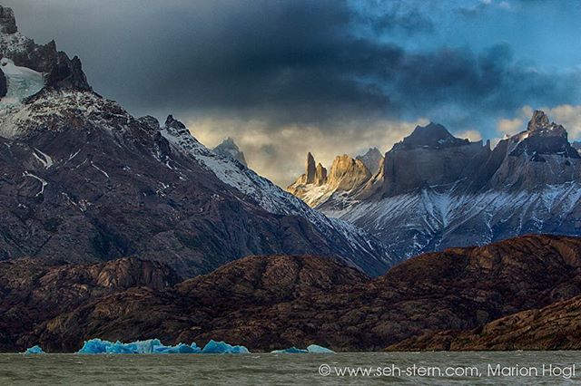 A majestic mountain range with snow-capped peaks and icebergs floating in a tranquil lake beneath a cloudy sky.