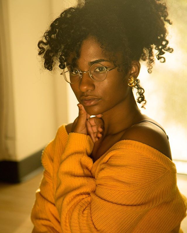 Portrait of a woman with curly hair wearing glasses and a yellow sweater.