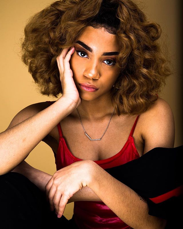 A woman with curly hair and a red top poses in a studio setting, offering a confident and sensual vibe.