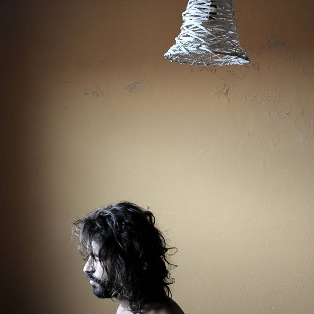 A man with long dark hair stands beneath a modern lamp in a moody, intimate portrait.