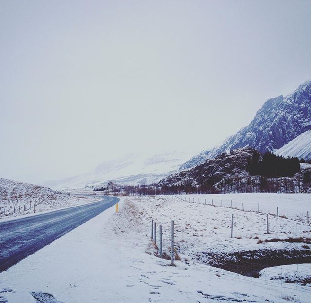 A snow-covered road winds through a serene winter landscape with mountains in the background.