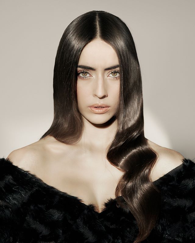 A brunette woman with sleek, long hair and fair skin poses in a studio setting for a beauty or fashion campaign.