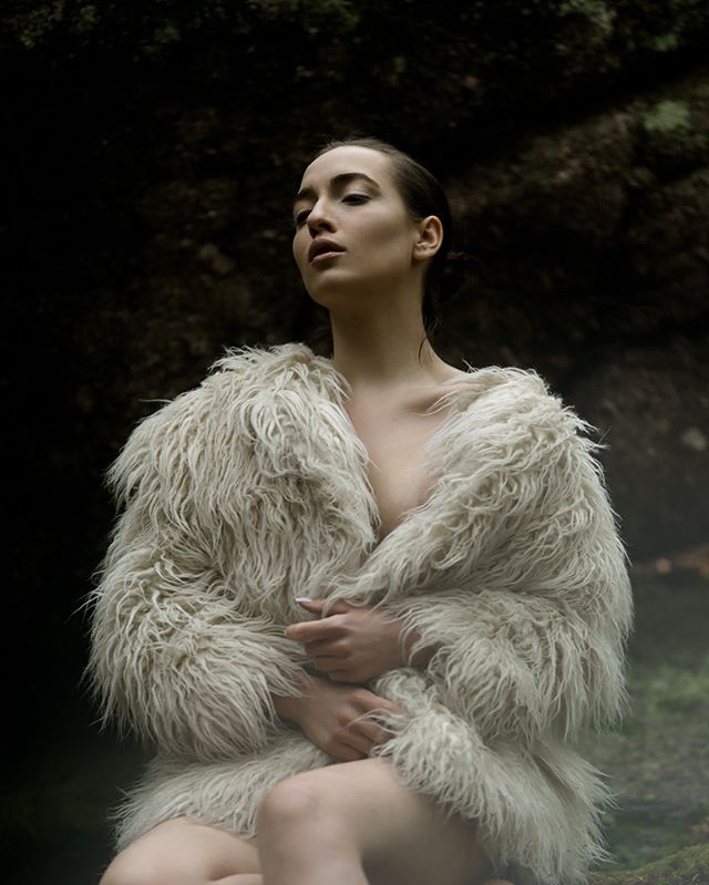 A woman in a fur coat poses outdoors in a soft, dreamy setting.