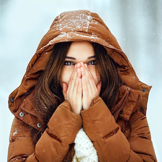 A brunette woman in a hooded winter jacket outside in the snow covers her face with her hands to stay warm.