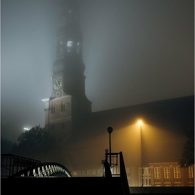 An atmospheric night scene featuring a clock tower shrouded in fog, illuminated by a street lamp and a bridge.