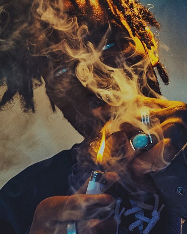 A man with dreadlocks lights up, surrounded by smoke. Moody and intense portrait.