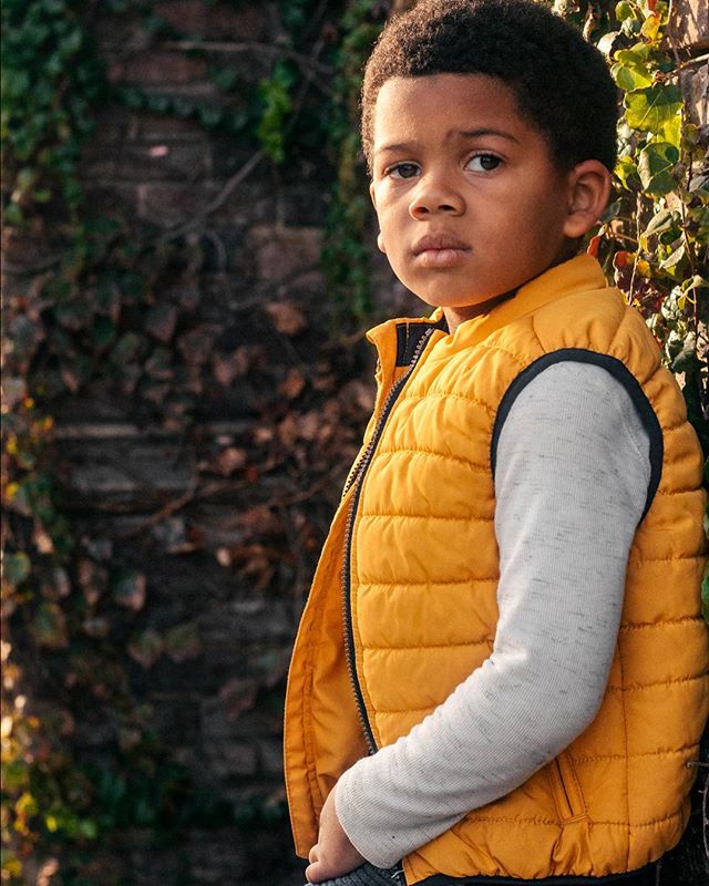 Portrait of a young boy in a yellow vest leaning against a vine-covered wall, looking thoughtfully at the camera.