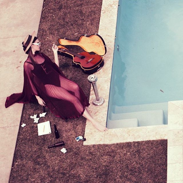 A woman lounges by a pool with a guitar and case, smoking a cigarette.