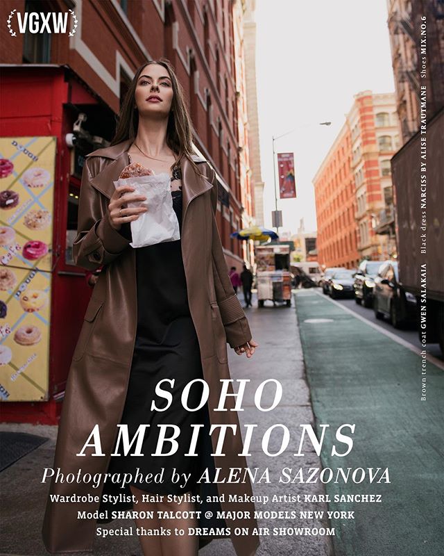 A fashionable woman enjoys a donut on a city street, wearing a trench coat and black dress for an editorial photoshoot.