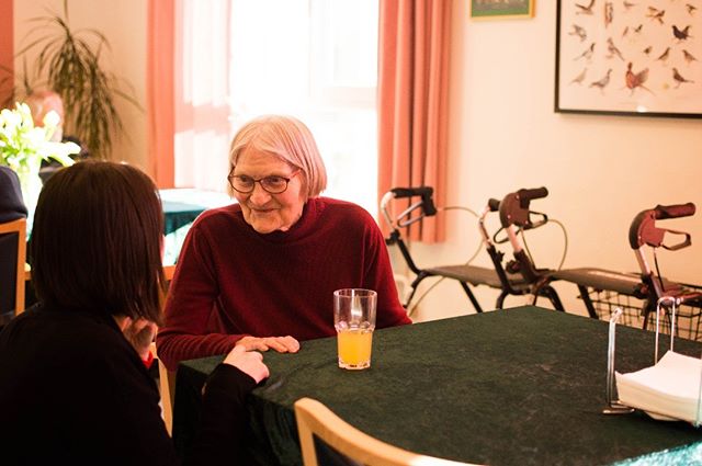 An elderly woman sits at a table and enjoys a glass of orange juice during a pleasant conversation.