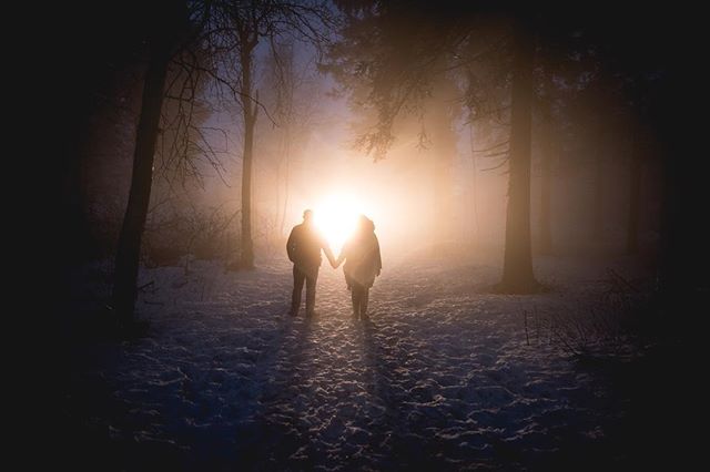 A couple walks hand in hand through a snowy forest, silhouetted against the bright sunlight.