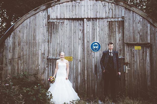 A bride and groom pose in front of a rustic wooden building on their wedding day.