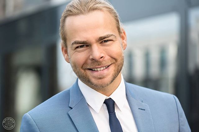 Confident businessman smiles wearing a blue suit and tie against a modern urban backdrop.