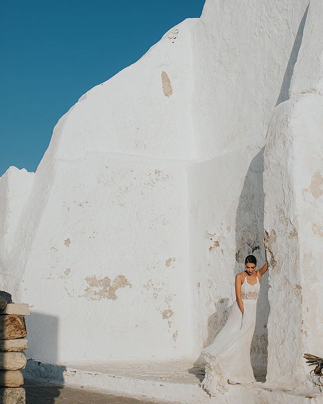 A woman in a white dress stands against a whitewashed building, bathed in soft light.