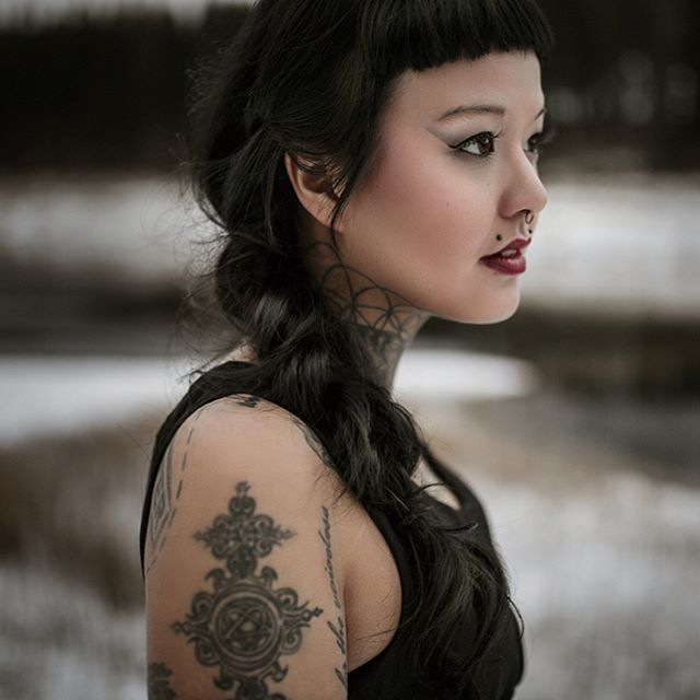 A tattooed woman with dark hair poses outdoors with a contemplative expression in a moody, editorial style photograph.