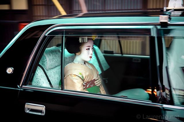 A geisha in traditional makeup sits in the back of a taxi, providing a glimpse into Japanese culture.