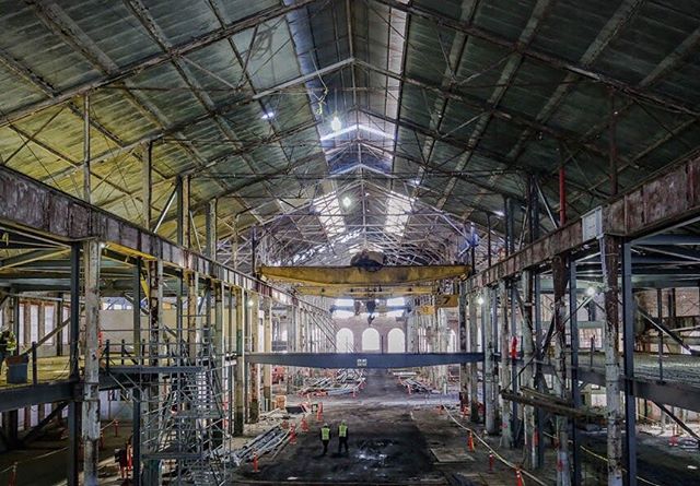 Interior view of an industrial building under construction, featuring scaffolding, crane, and workers in a vast, open space.