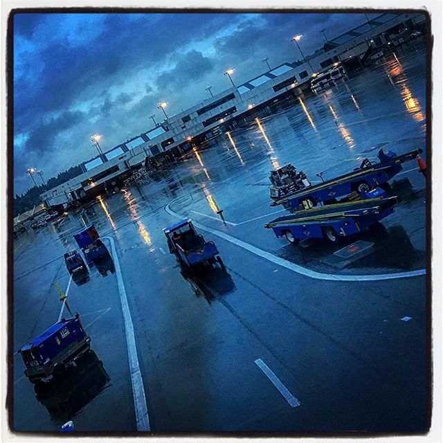 Airport ground vehicles and luggage tugs sit on the wet tarmac at night, with lights reflecting on the surface.