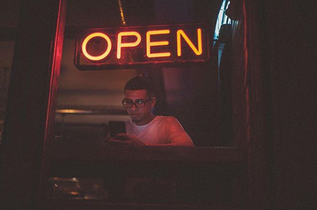 A man looks at his phone underneath an illuminated 'Open' sign, casting a warm glow.