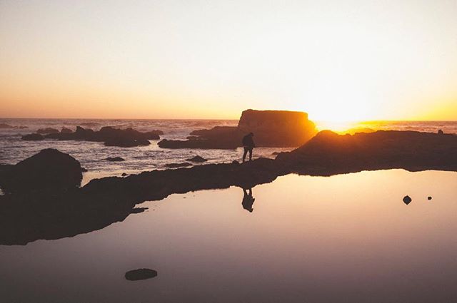 A lone person walks along a rocky coastline at sunset, their silhouette reflected in a tide pool.