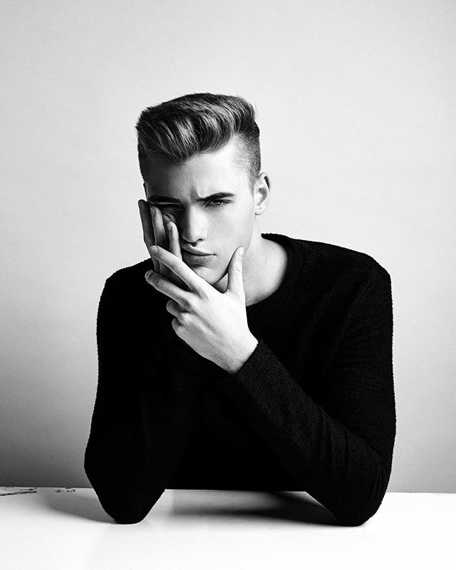 A black and white portrait of a serious young man in a black sweater, thoughtfully posing in a studio.