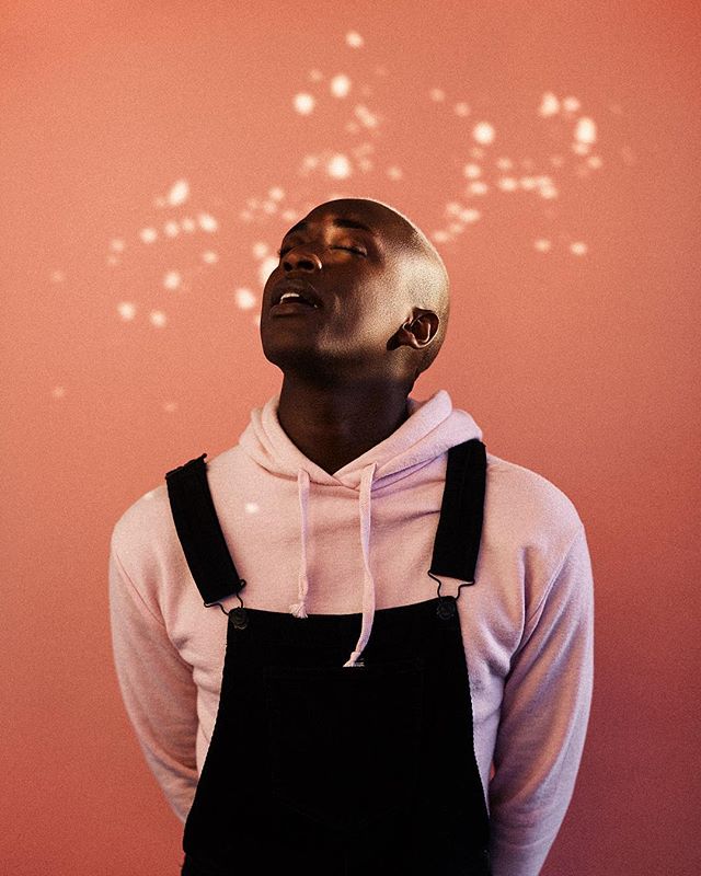 A bald African American man with eyes closed wearing a pink hoodie and black overalls, standing in front of a pink wall.