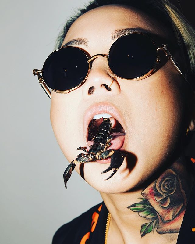 A woman with sunglasses has a scorpion in her mouth. The image is modern, striking, and attention-grabbing.