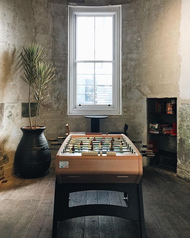 A rustic room features a foosball table, a plant, a window, and a bookshelf.