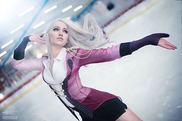 A cosplayer dressed in an elaborate figure skating costume poses gracefully on an ice rink with soft lighting.
